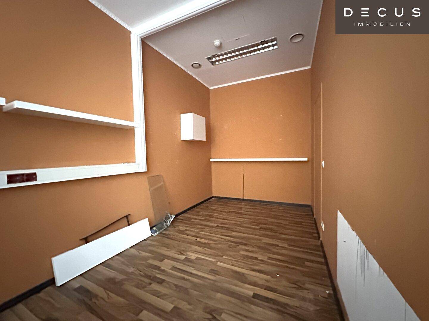 Kleiner Büroraum mit orangefarbenen Wänden, Laminatboden und einfachen weißen Regalen.