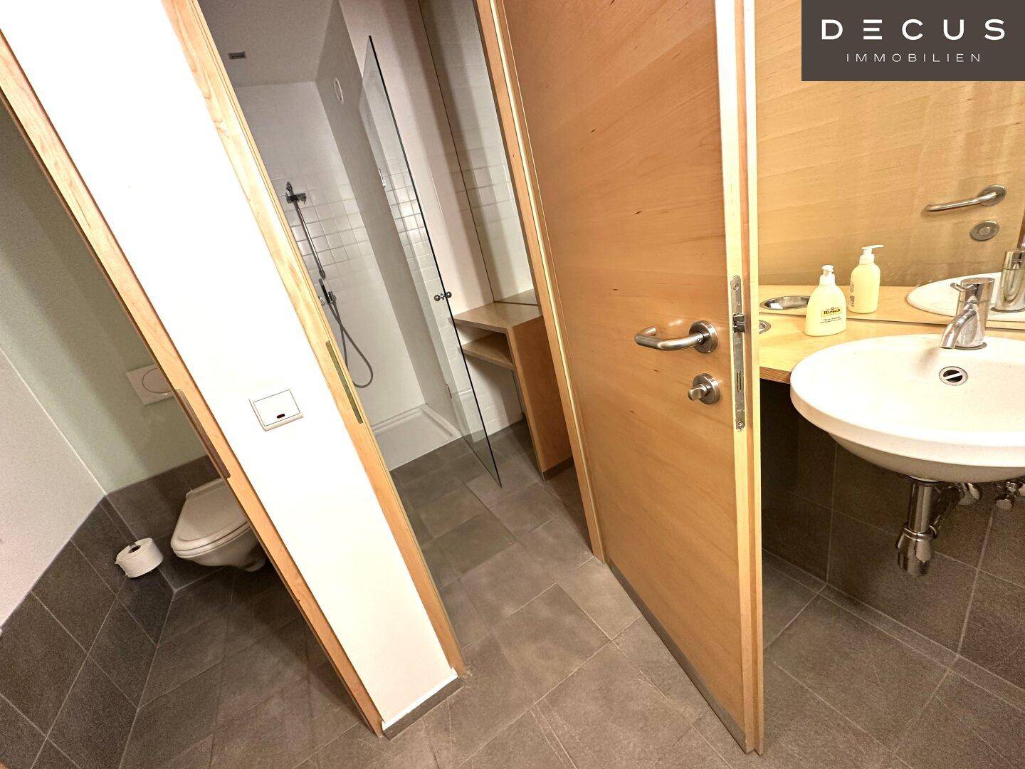 Badezimmer mit Toilette, Waschbecken und einer Dusche, die durch eine offene Tür sichtbar ist.