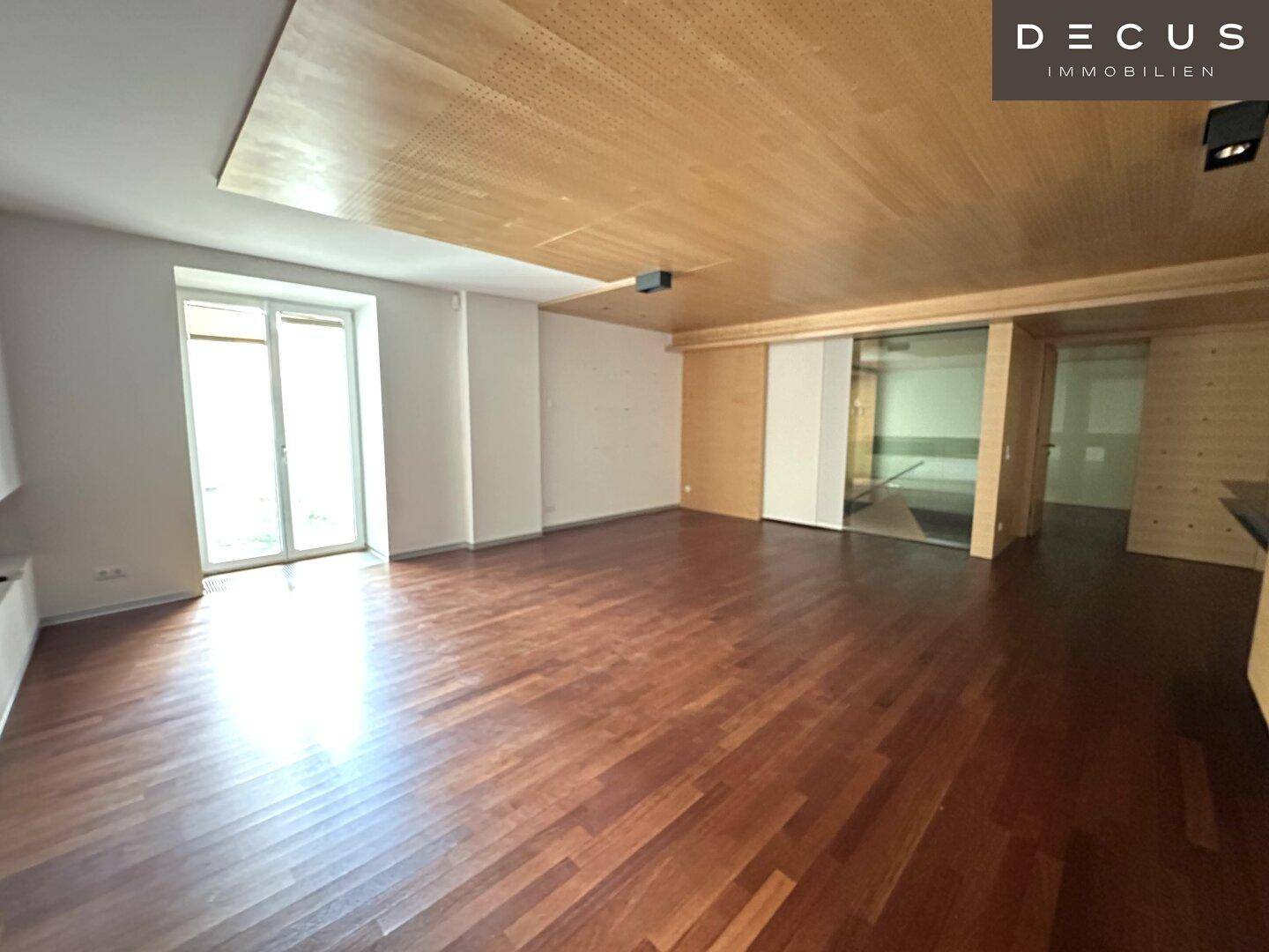 Heller, offener Wohnbereich mit Holzboden und Holzlamellendecke, große Fenster sorgen für viel Licht.