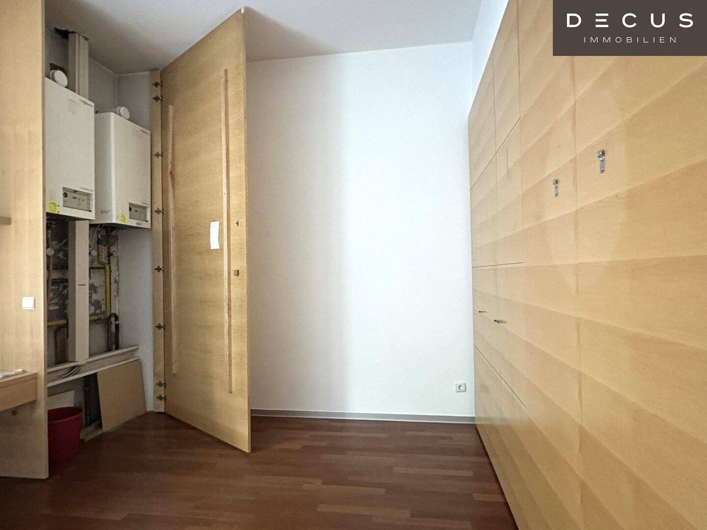 Raum mit Holzboden und Holzvertäfelung, eine offene Tür zeigt den Technikraum mit Heizkesseln.