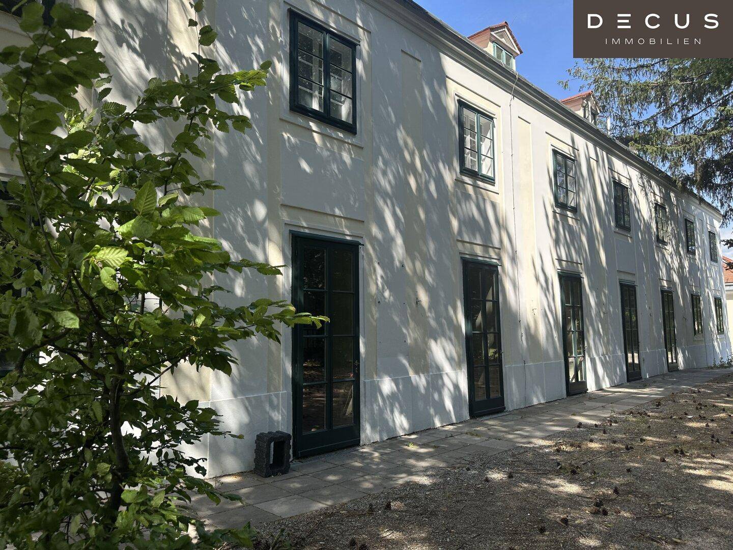 Lange Fassade eines historischen Gebäudes mit vielen Fenstern und Türen, umgeben von Grün.