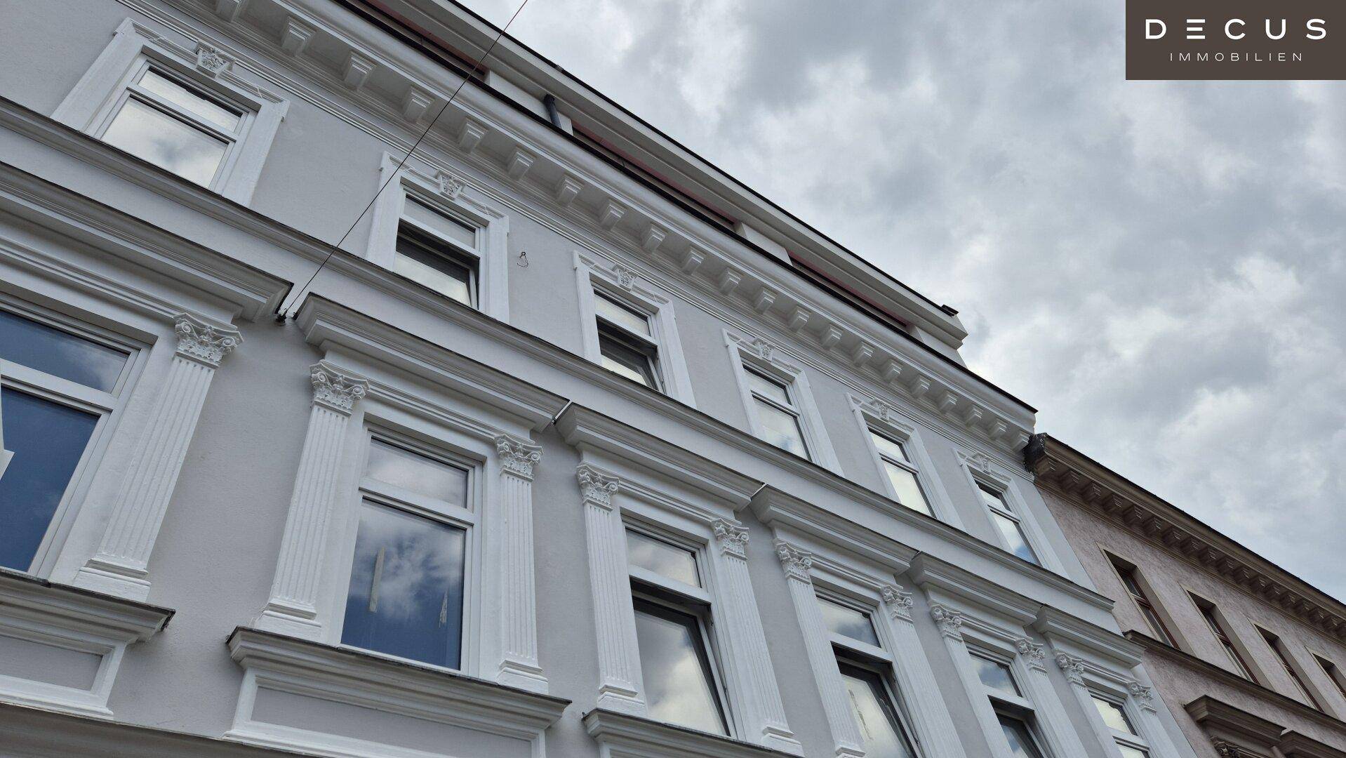 Klassische Gebäudefassade mit dekorativen Elementen und großen Fenstern unter einem bewölkten Himmel.