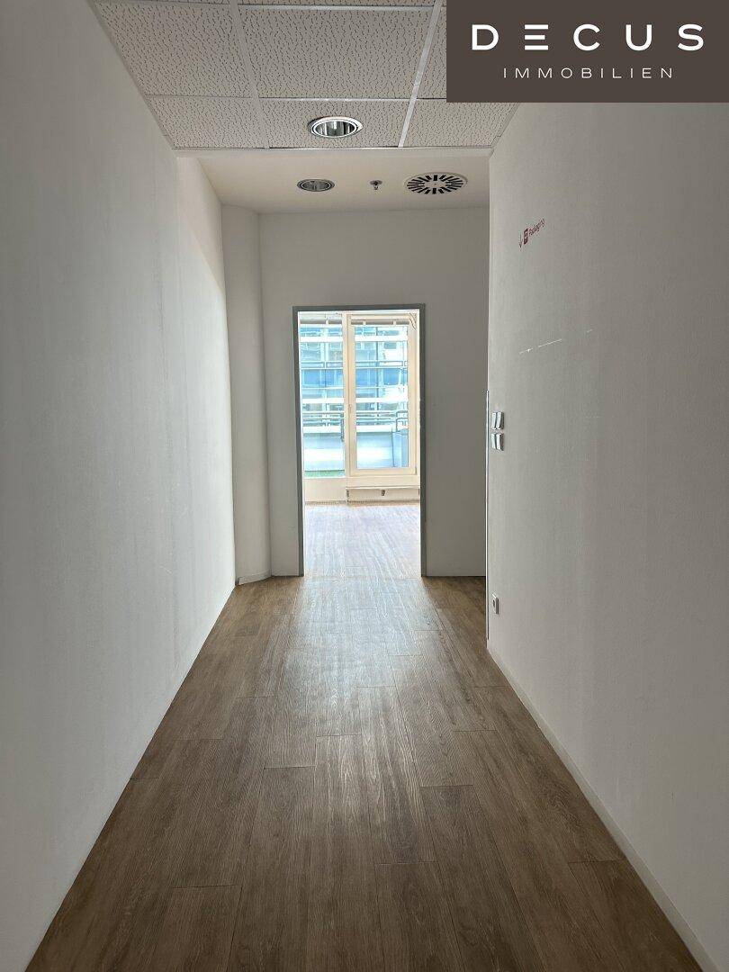 Leerer Büroflur mit Laminatboden und weißen Wänden, der zu einem hellen Raum mit Fenstern führt.