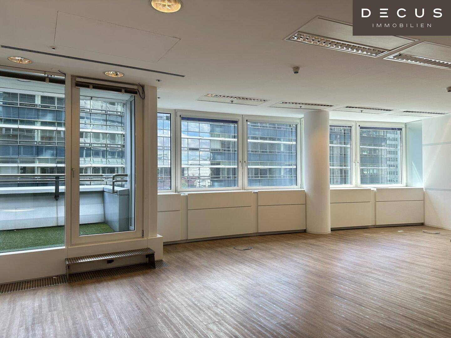 Geräumiger, leerer Büroraum mit großen Fenstern, Laminatboden und Zugang zu einer kleinen Terrasse.