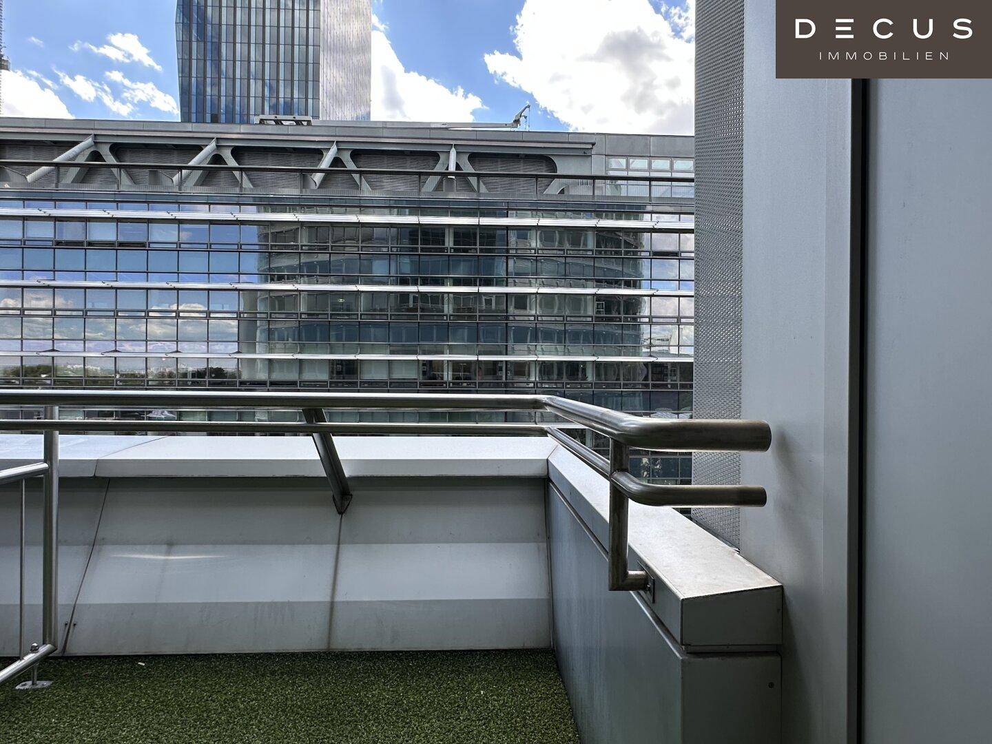 Kleiner Balkon mit Kunstrasen und Blick auf moderne Bürogebäude unter blauem Himmel.