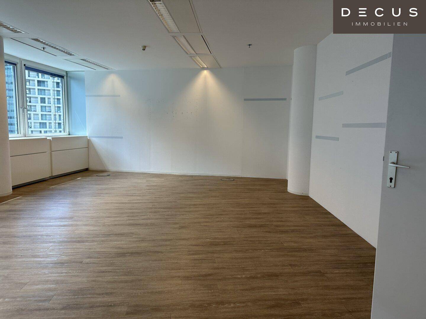 Leerer Büroraum mit Laminatboden und weißen Wänden, beleuchtet durch Fenster an einer Seite.
