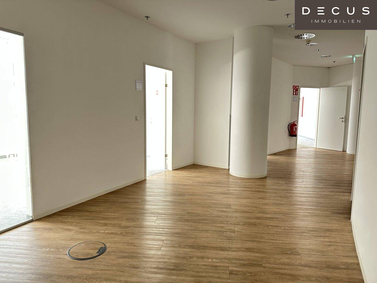 Geräumiger, leerer Bürobereich mit Laminatboden, einer runden Säule und mehreren Türen zu weiteren Räumen.