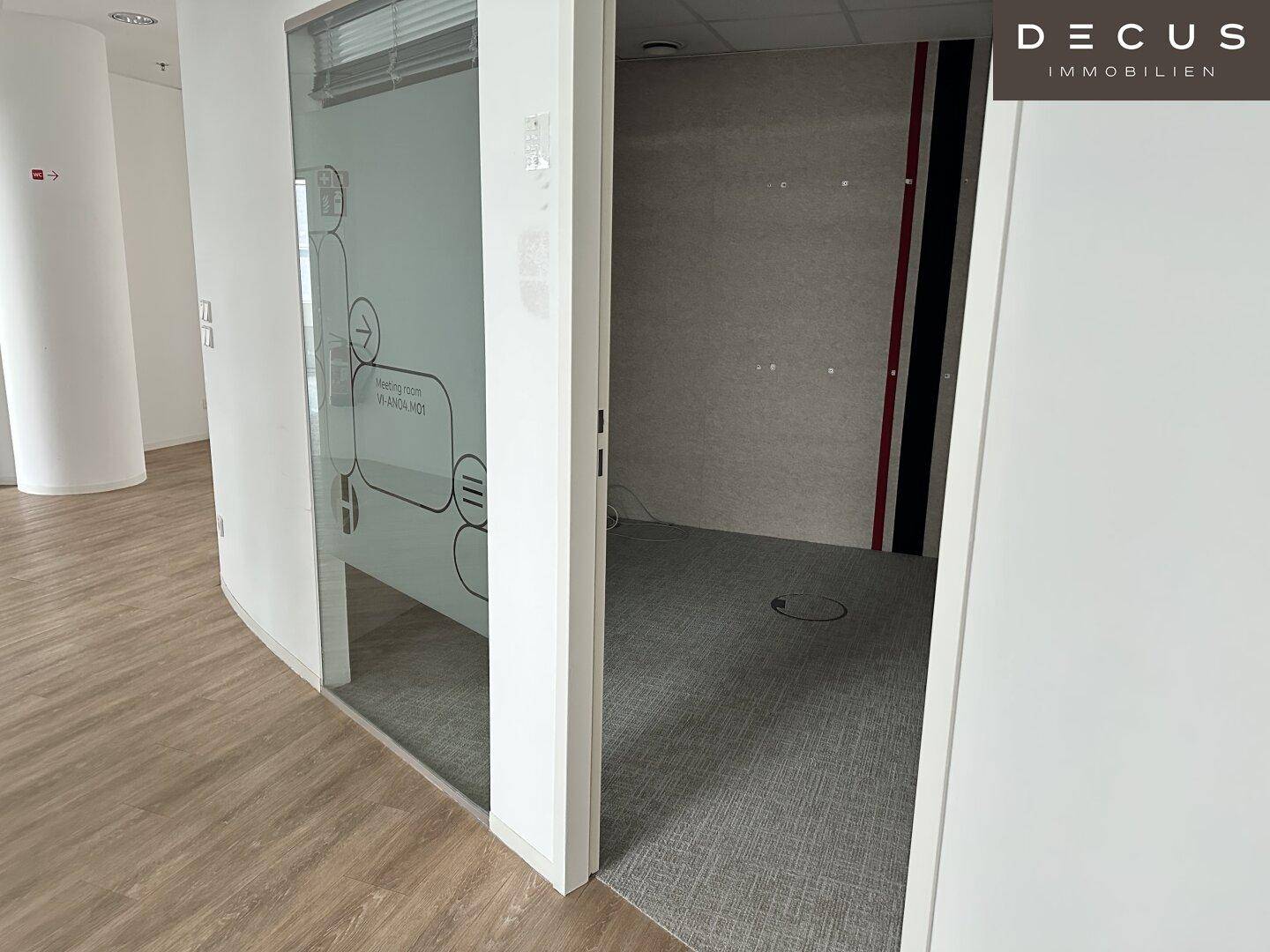 Büro mit Glaswand, die zu einem kleineren Raum mit Teppichboden führt, ideal für Besprechungen.