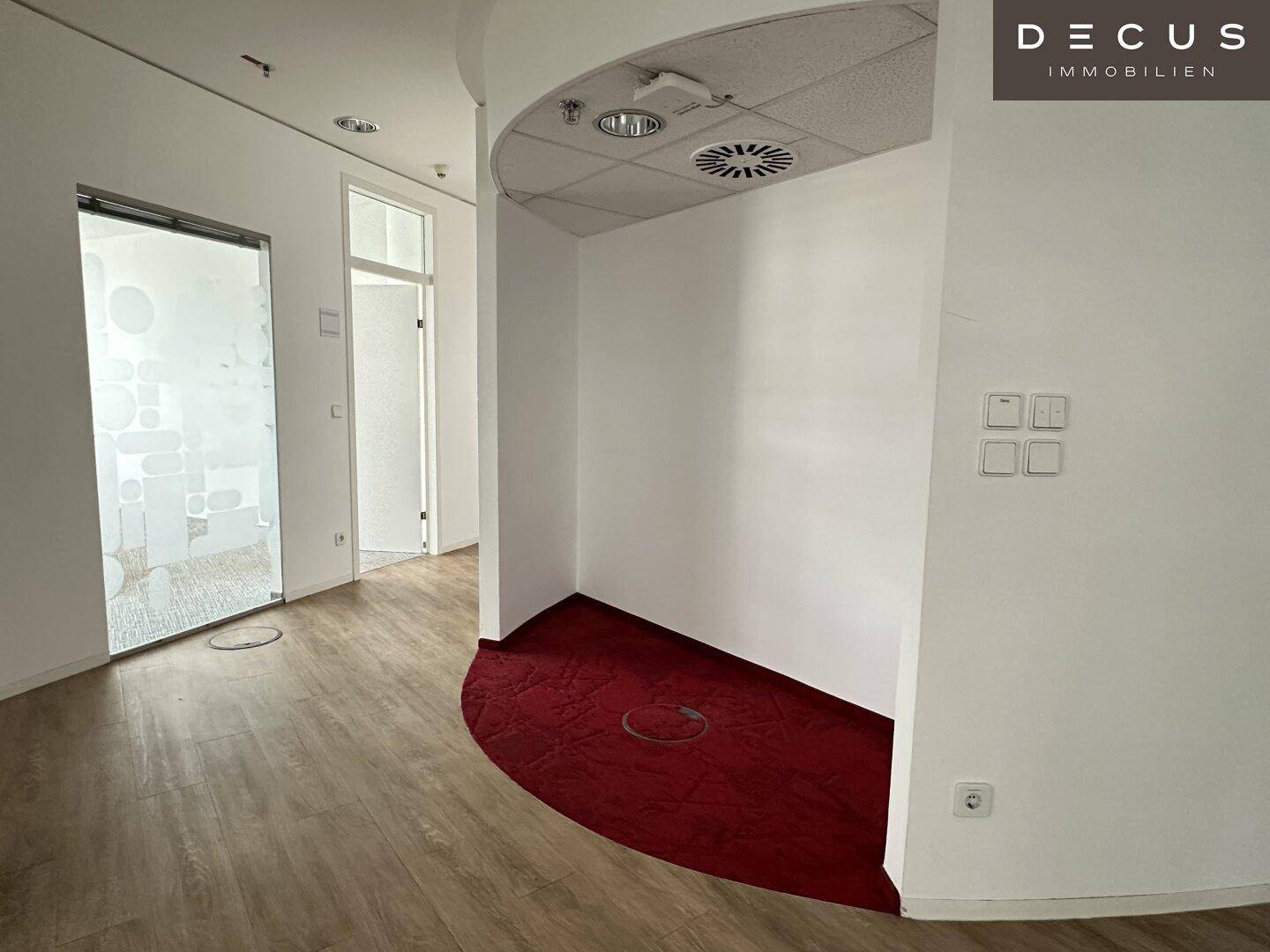 Bürobereich mit geschwungener Wand, Laminatboden und einem Akzentbereich mit rotem Teppich.