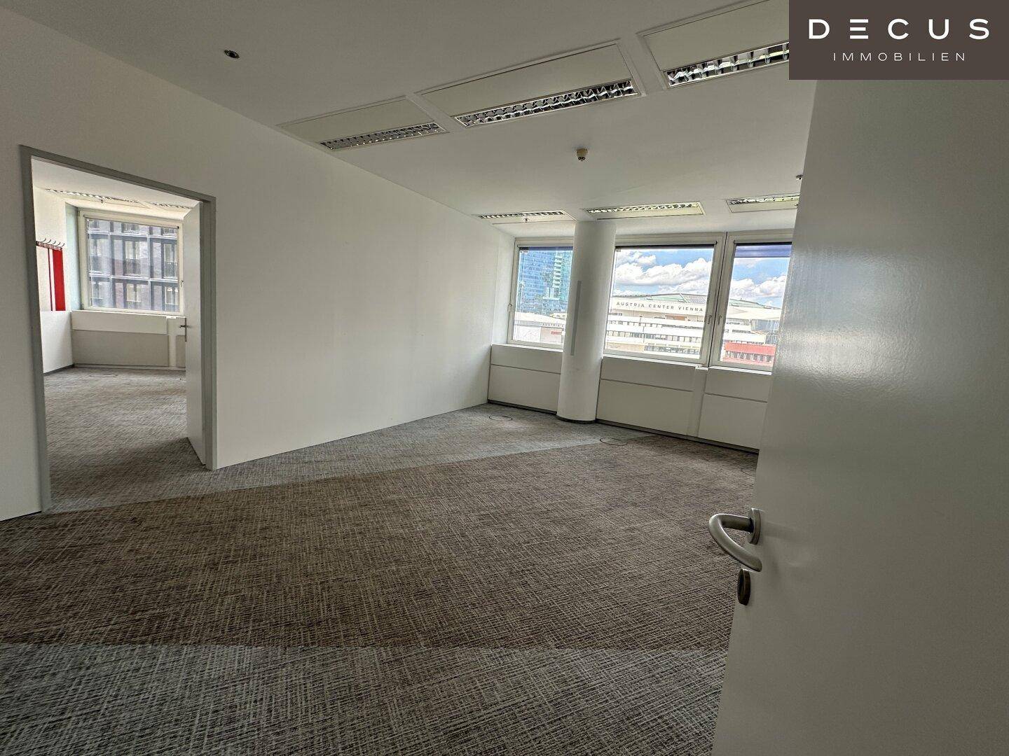 Leerer Büroraum mit Teppichboden und Fenstern, die Tageslicht hereinlassen, gesehen durch eine offene Tür.