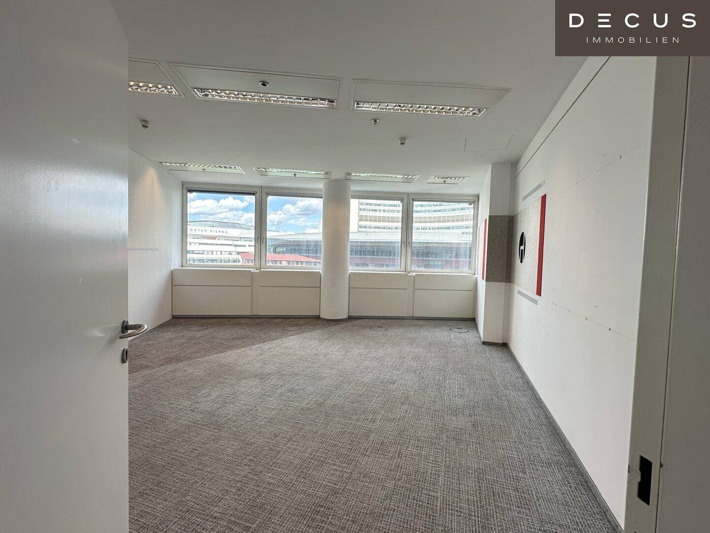 Heller, leerer Büroraum mit Teppichboden und Fenstern, die einen Blick nach draußen ermöglichen.