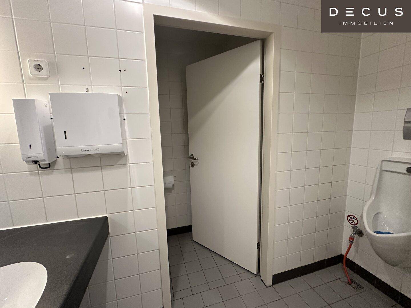 Öffentliche Toilette mit weißen Fliesen, einem Urinal und einem Waschbeckenbereich mit Spender.