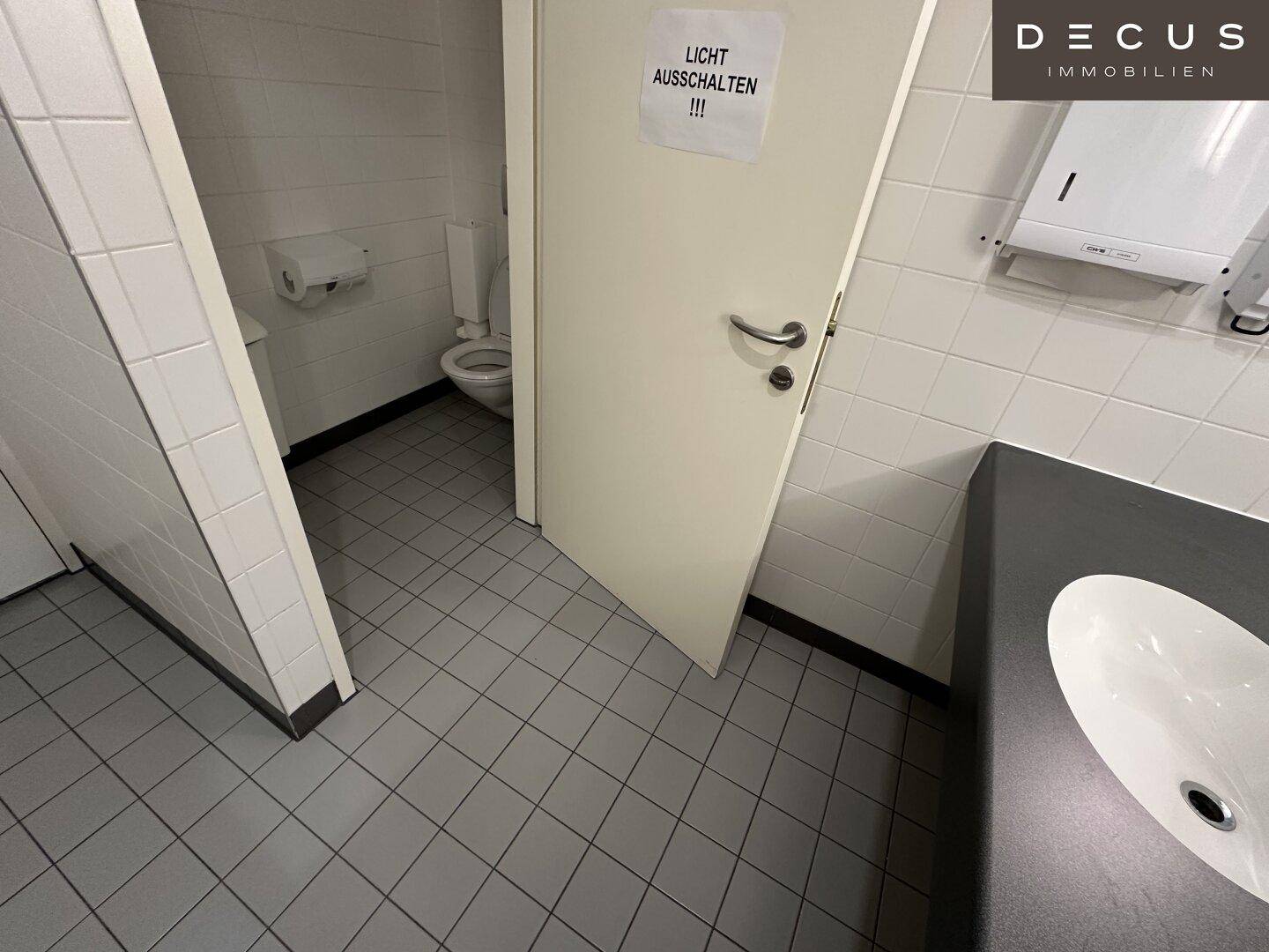 Saubere öffentliche Toilette mit weißen Fliesen, WC und Waschbecken, mit Hinweis zum Lichtausschalten.