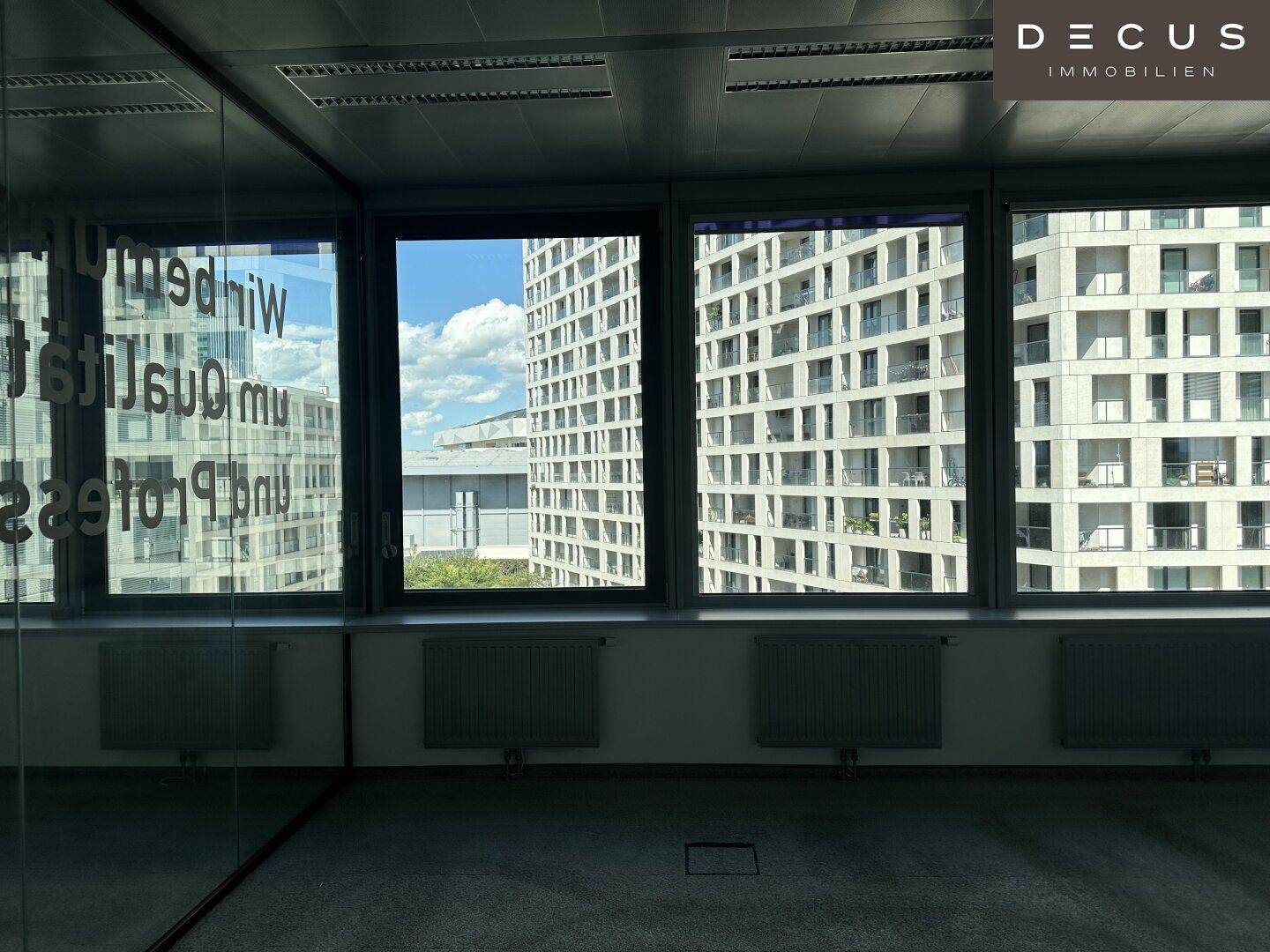 Bürofenster mit Blick auf eine moderne Wohnanlage und den blauen Himmel.