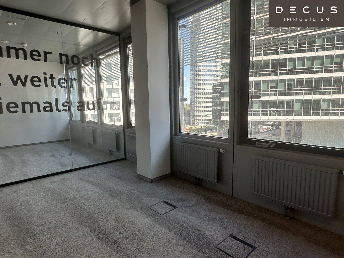 Helles Büro mit Teppichboden und großen Fenstern, die einen Blick auf andere Gebäude bieten.
