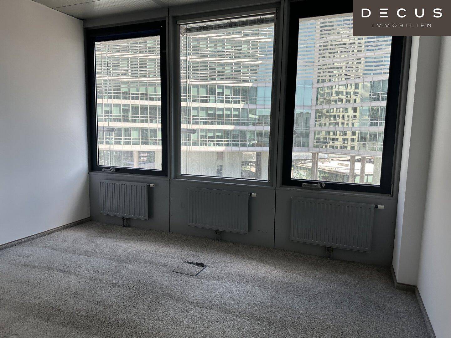 Kleines Büro mit Teppichboden und großen Fenstern, die einen weiten Blick auf die Stadt ermöglichen.