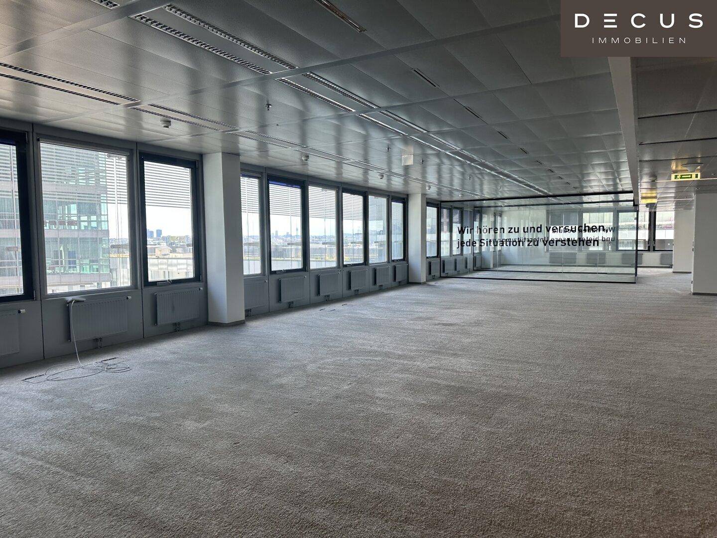 Großes, offenes Büro mit Teppichboden, Glaswänden und einer langen Fensterfront mit Stadtblick.