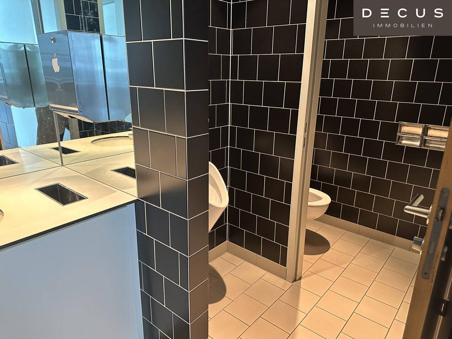 Moderne Herrentoilette mit schwarzen Fliesen, Urinal und Waschbeckenbereich mit Spiegel.