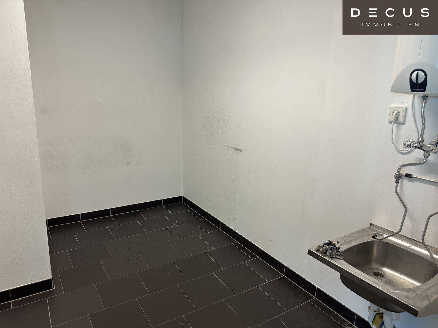 Kleiner Abstellraum mit Waschbecken, Boiler und dunklen Fliesen am Boden.