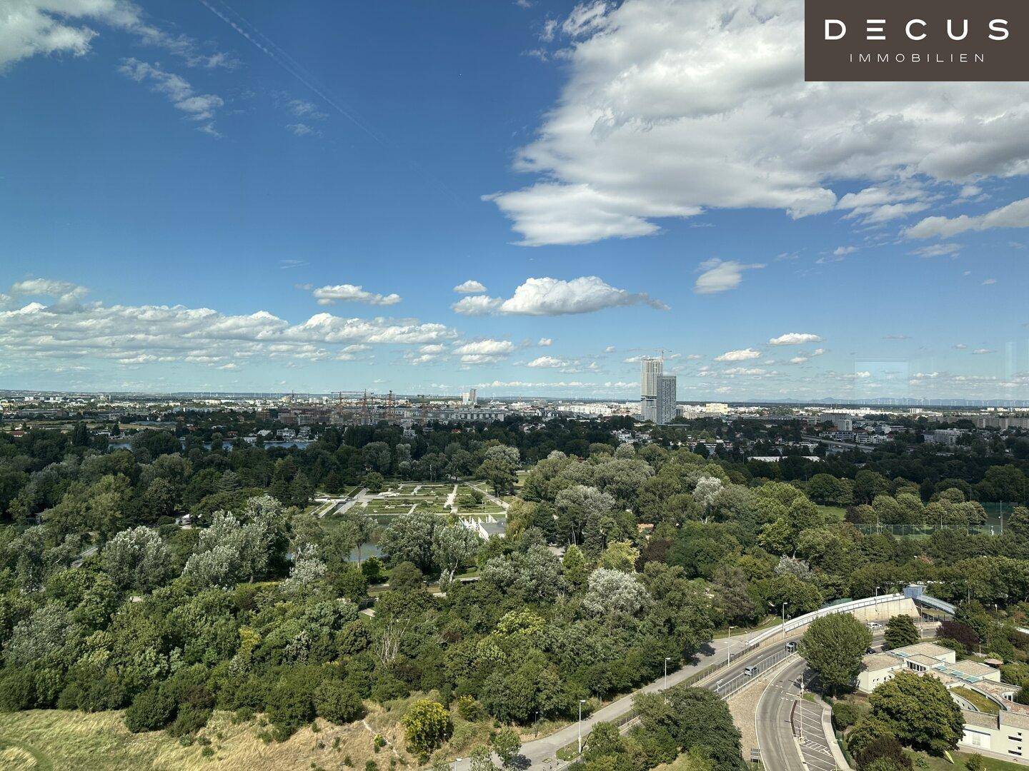 Weitläufiger Blick über eine grüne Landschaft mit Park, See und städtischer Skyline am Horizont.