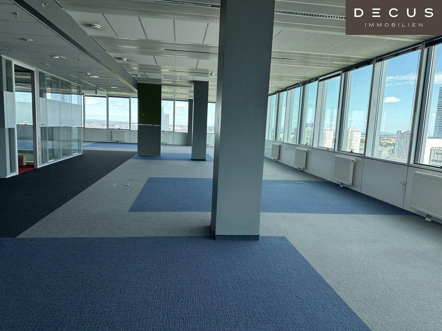 Offener Bürobereich mit Säulen, Teppichboden und raumhohen Fenstern mit Stadtblick.