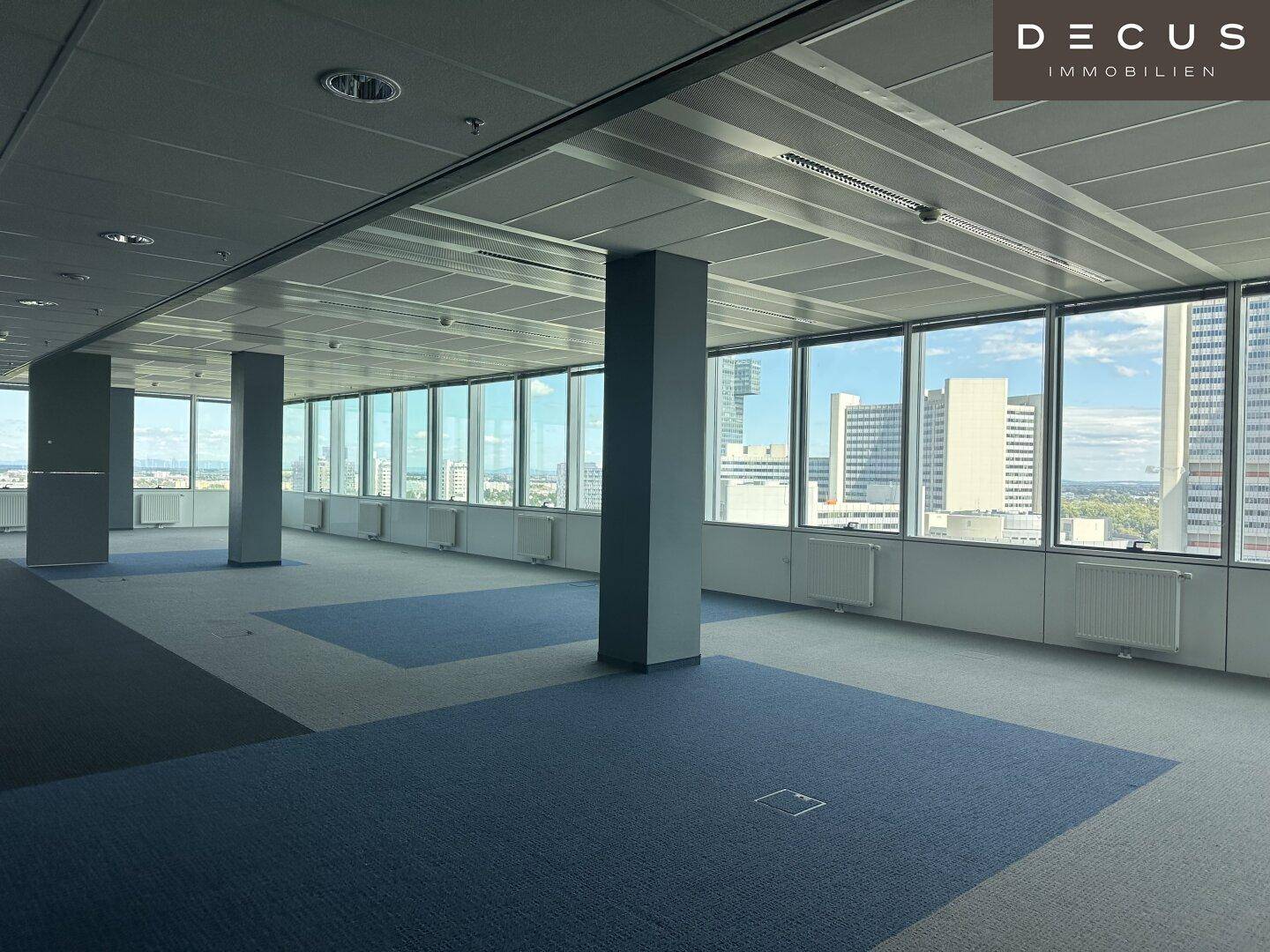 Großzügiger Büroraum mit Teppichboden, hohen Decken und Panoramablick auf die Stadt.