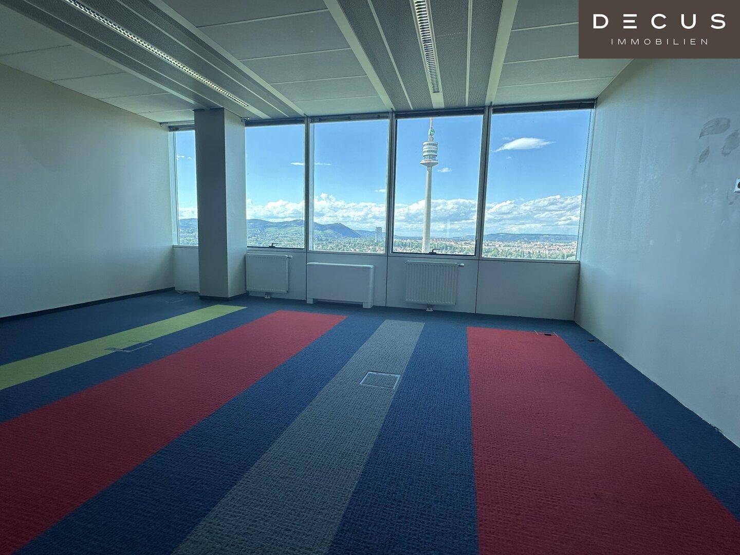 Großes Büro mit farbenfrohem Teppichboden und weitem Ausblick auf die Stadt und den Donauturm.