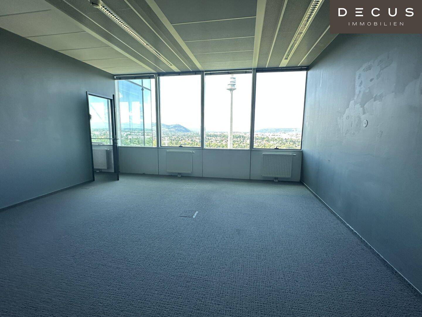Leeres Büro mit Teppichboden und großen Fenstern, die einen weiten Blick auf die Umgebung bieten.