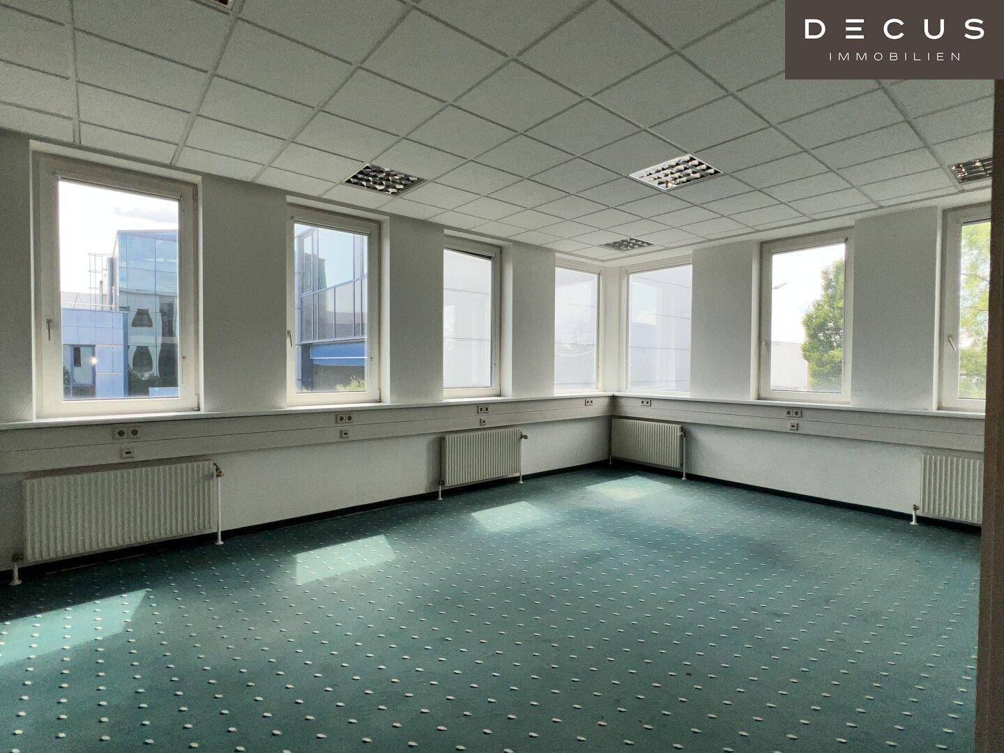 Helles Eckbüro mit grünem Teppichboden, vielen Fenstern und einer praktischen Deckenbeleuchtung.