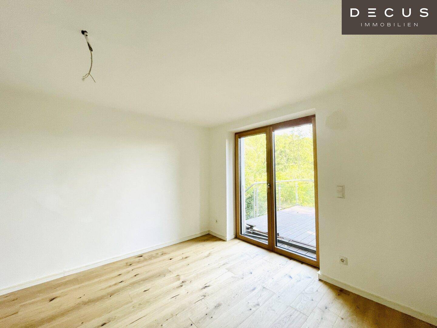 Heller Raum mit neuem Holzboden und großem Fenster mit Zugang zu einem Balkon und Blick ins Grüne.