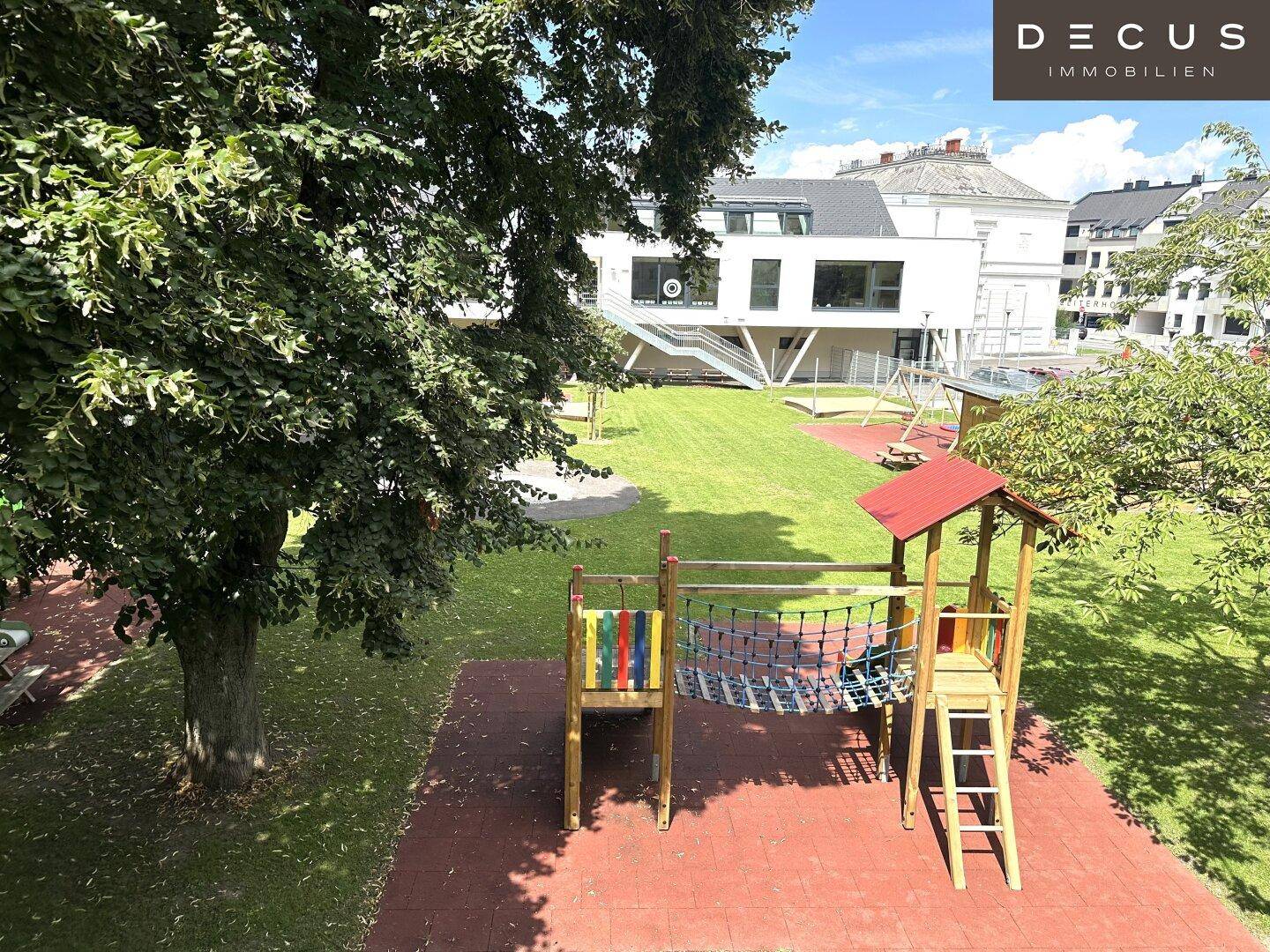 Kinderspielplatz mit Schaukeln und Klettergerüst, umgeben von Bäumen und Grünflächen.