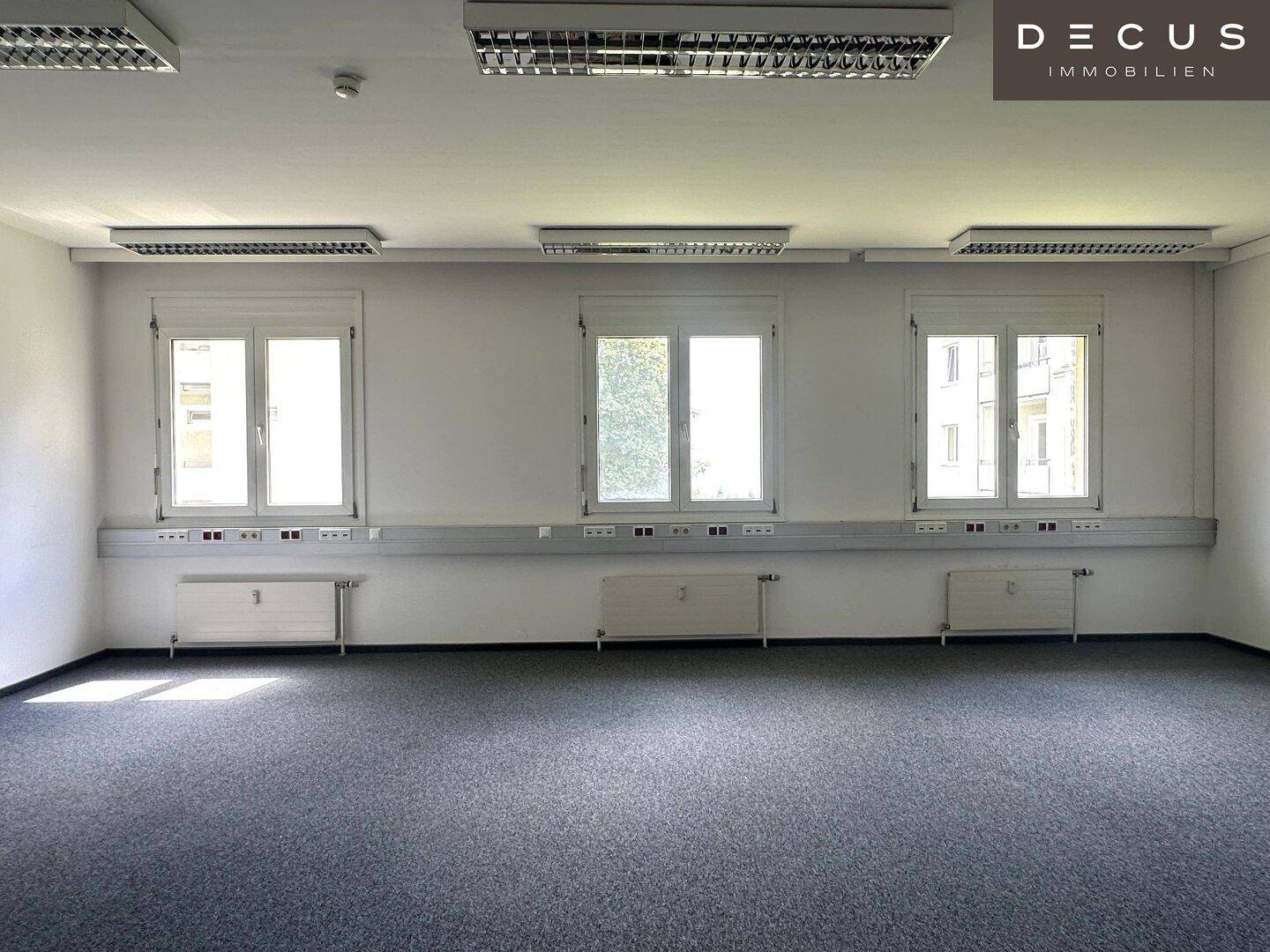 Geräumiges, leeres Büro mit drei Fenstern, Teppichboden und Deckenbeleuchtung.