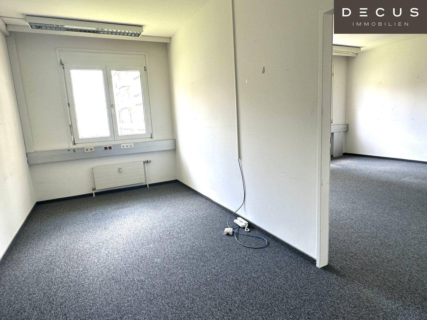 Helles Büro mit Teppichboden, einem Fenster und Zugang zu einem weiteren Raum.