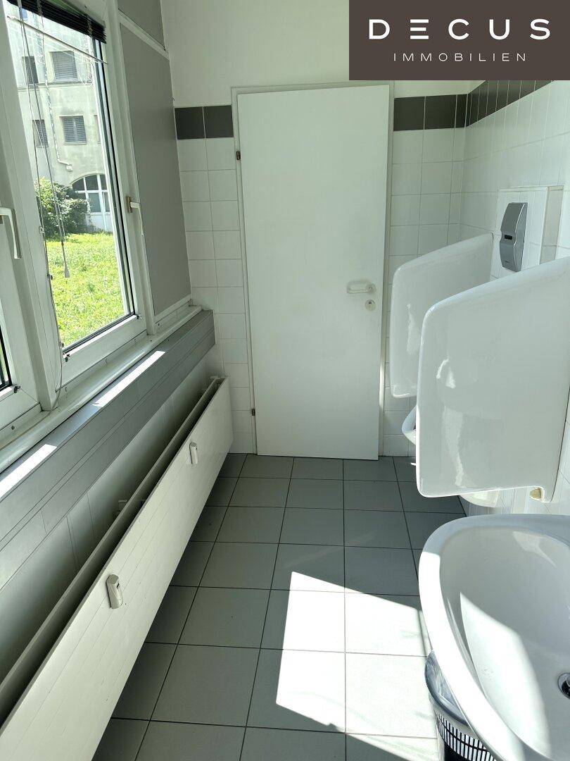 Herren-WC mit Urinalen, Waschbecken und Fenster für natürliche Belüftung und Tageslicht.