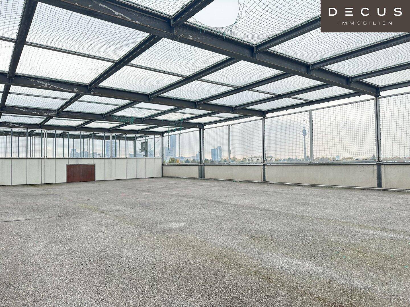 Große, leere Betonfläche auf einem Parkdeck mit Gitterdach und Blick auf die Stadt im Hintergrund.