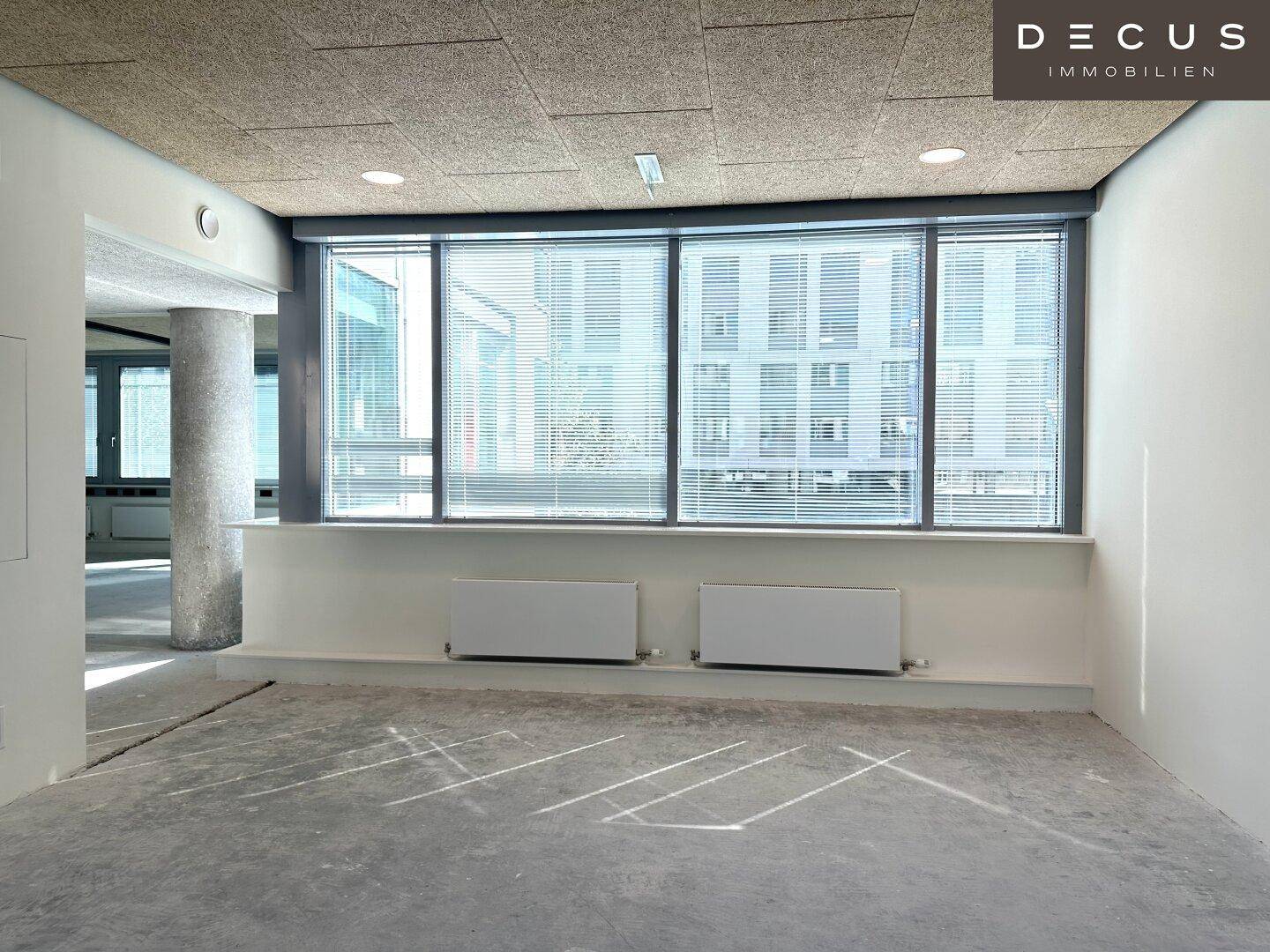 Unmöblierter Büroraum mit großen Fenstern, Heizkörpern und einer strukturierten Decke.