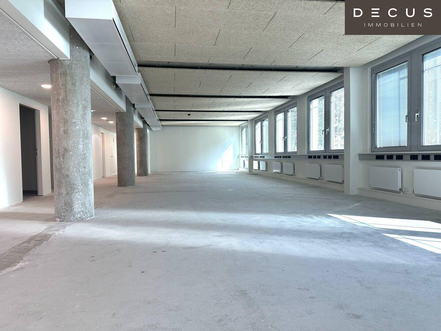 Langer, unmöblierter Büroraum mit vielen Fenstern, Säulen und einer Akustikdecke.