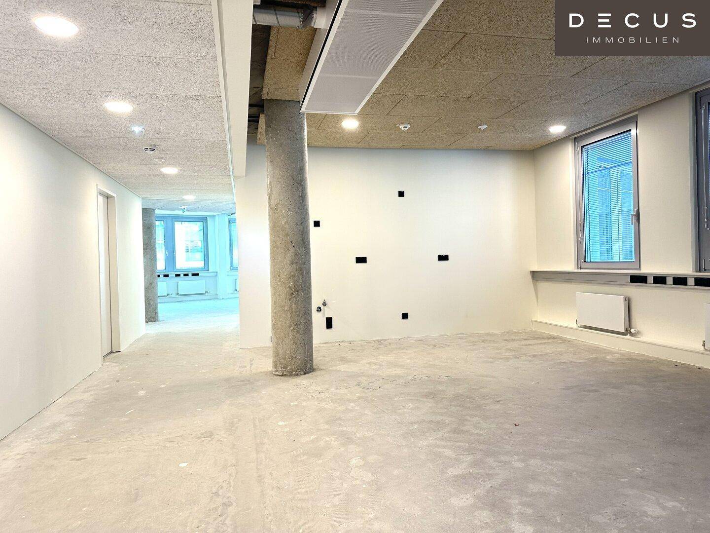 Unmöblierter Büroraum mit einer Säule, vielen Steckdosen und einer Akustikdecke.