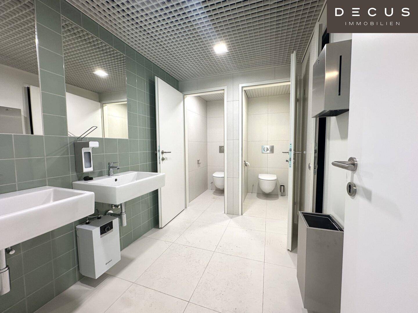 Moderner Waschraum mit zwei Waschbecken, Spiegeln und Toilettenkabinen im Hintergrund.