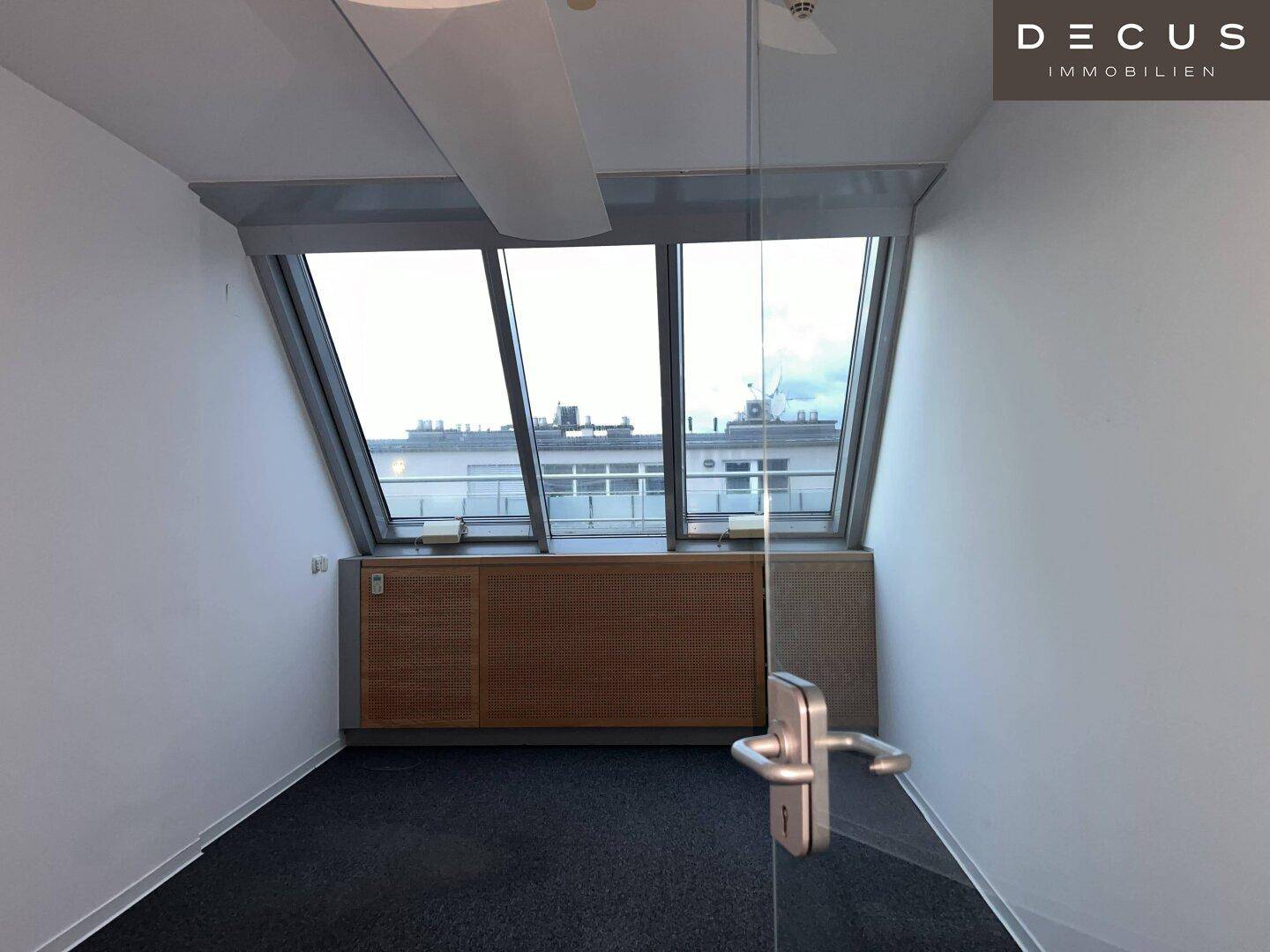 Helles Büro mit schrägen Fenstern und Einbauschränken, durch eine moderne Glastür zugänglich, mit Stadtblick.