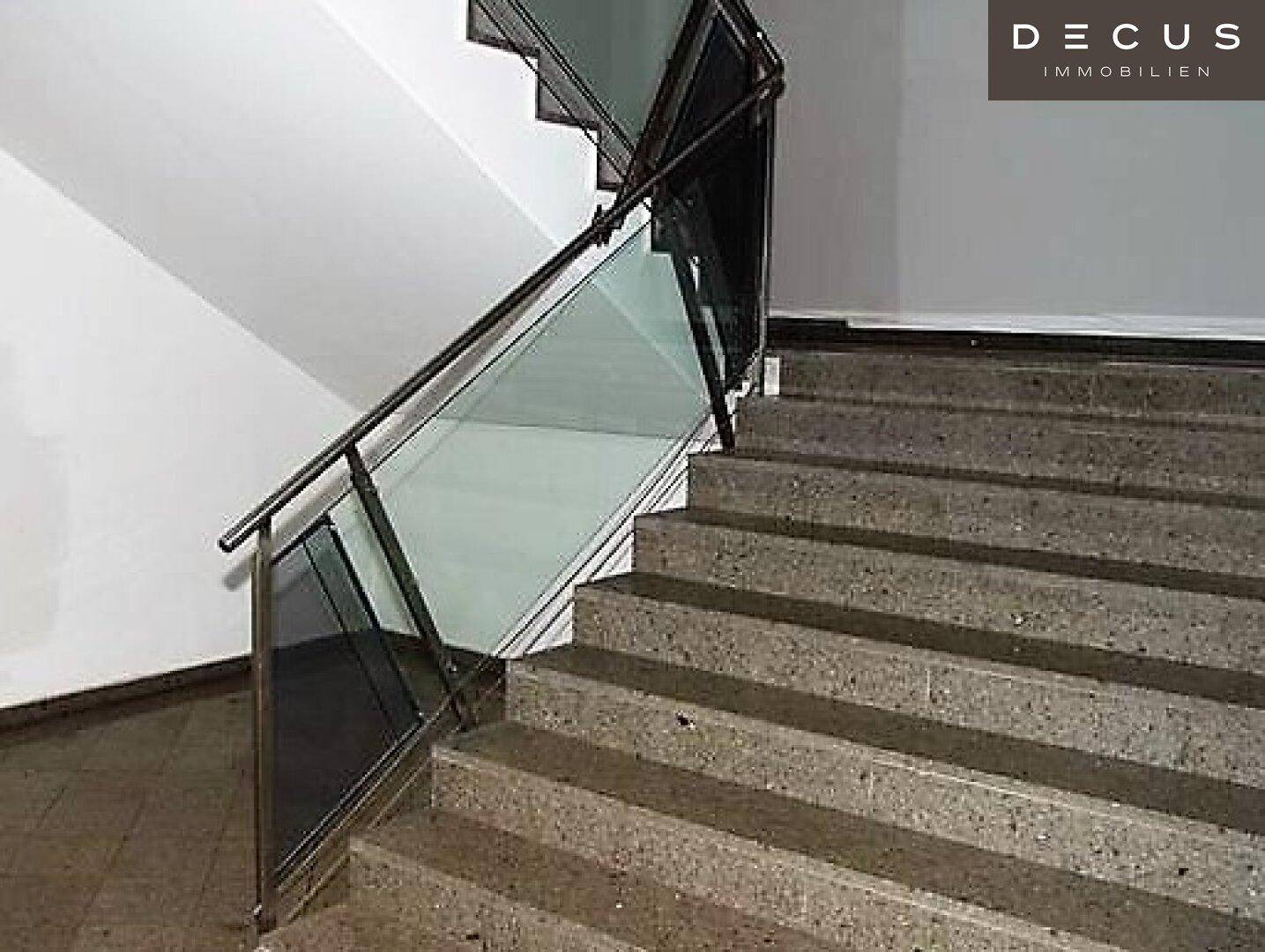 Helles Treppenhaus mit robusten Betonstufen und einem modernen Geländer aus Glas und Metall.