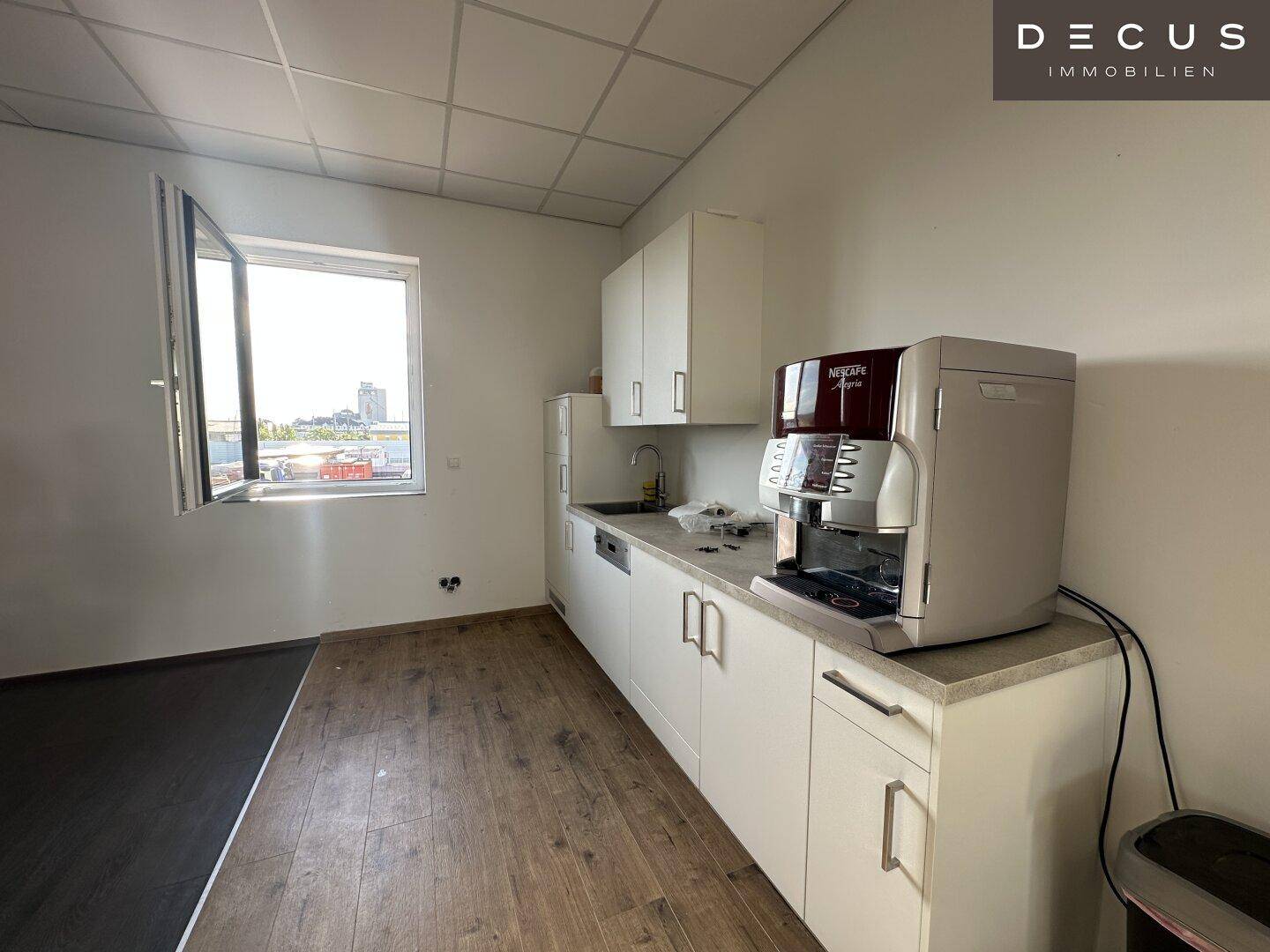 Funktionale Küchenzeile in einem Büroraum mit Fenster, Kaffeemaschine und integrierten Geräten.