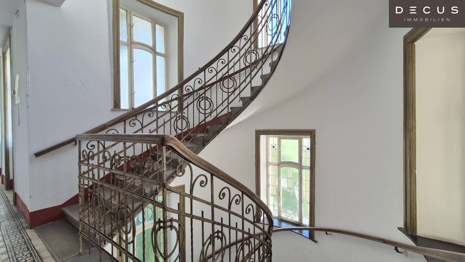Elegantes Treppenhaus mit geschwungener schmiedeeiserner Balustrade und großen Fenstern, typisch für Altbauten.