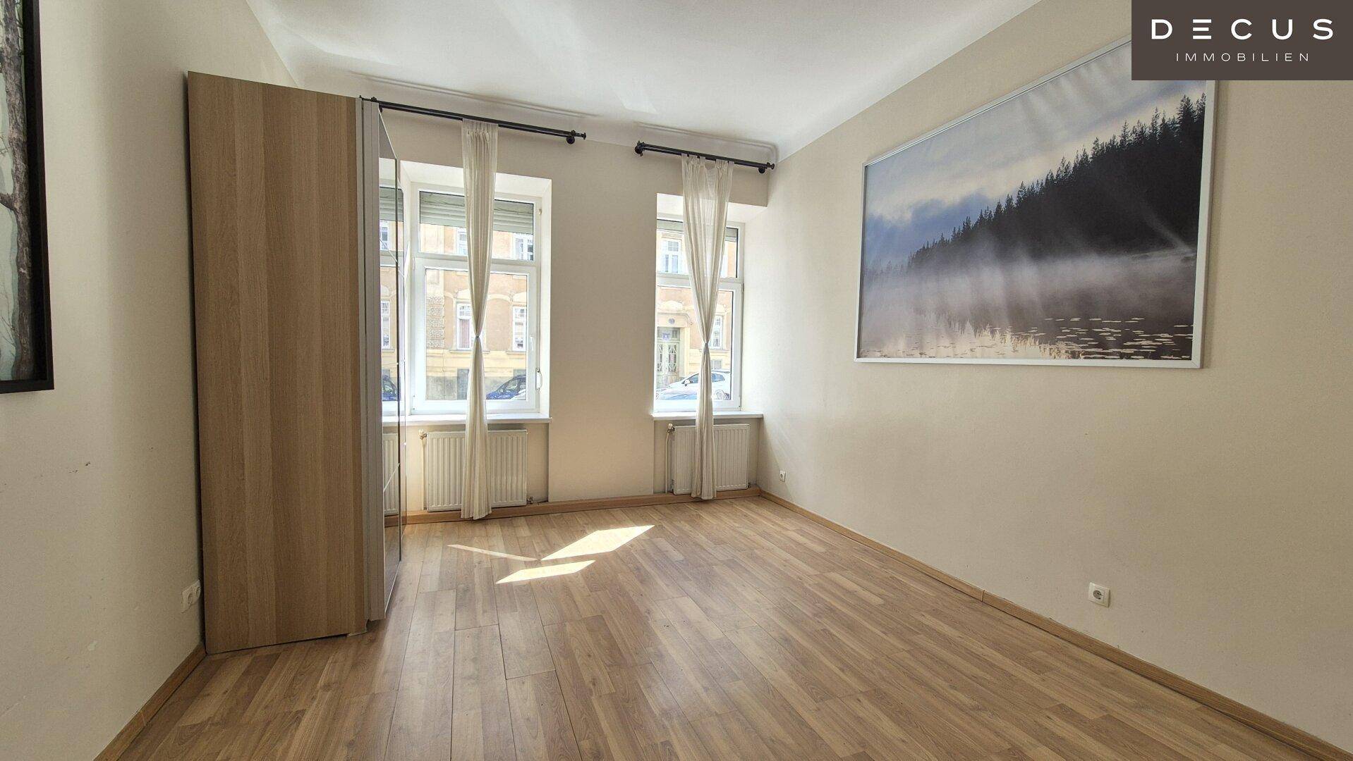 Helles Zimmer mit Holzboden, zwei Fenstern und einem großen Wandbild an der Wand.