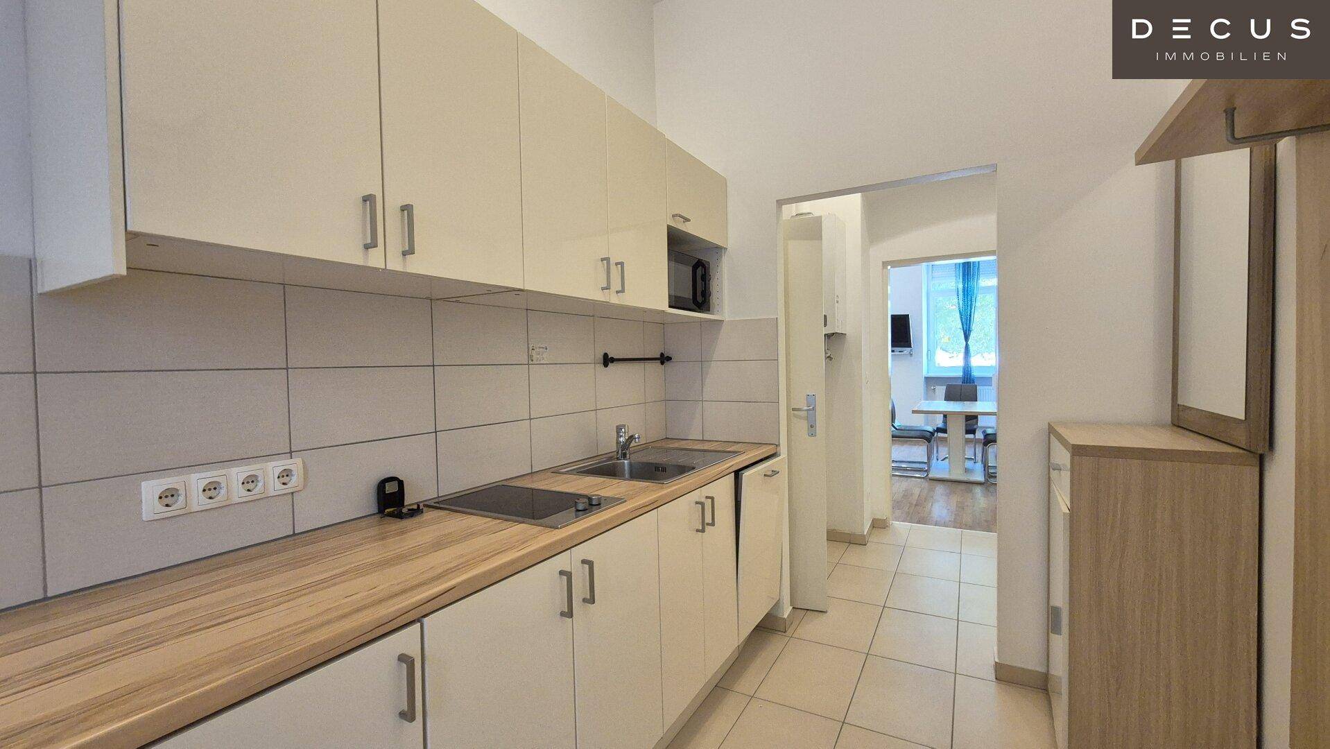 Funktionale Küche mit hellen Schränken, Holz-Arbeitsplatte und Fliesenboden, offen zum Eingangsbereich.