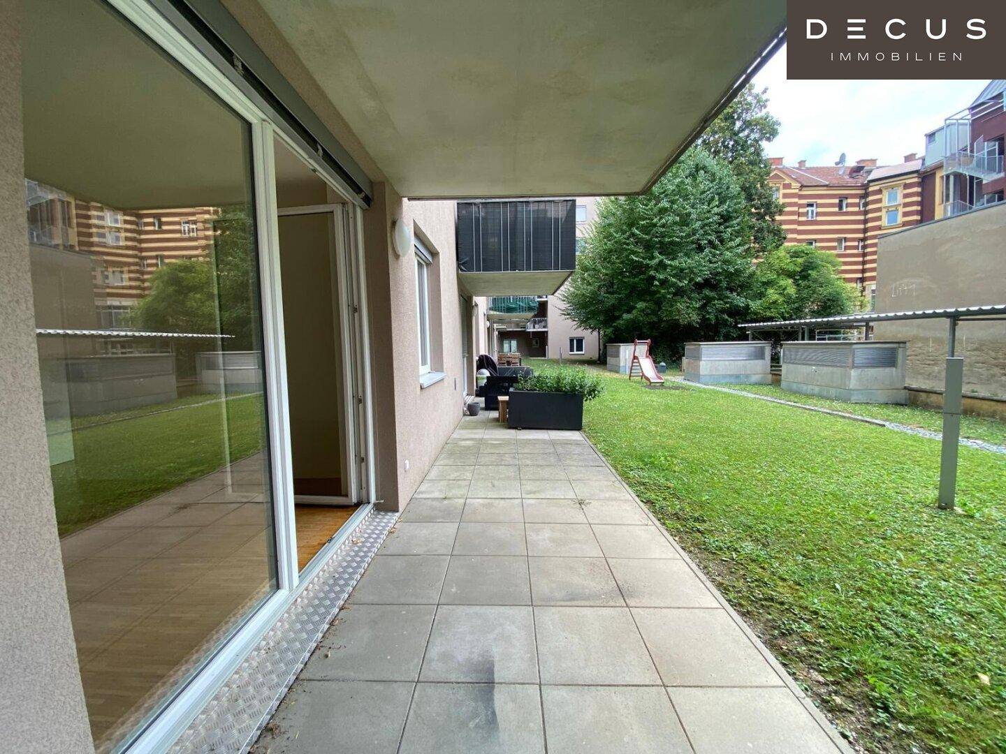 Gepflasterte Terrasse mit Zugang zu einem Gemeinschaftsgarten und einem kleinen Spielbereich.