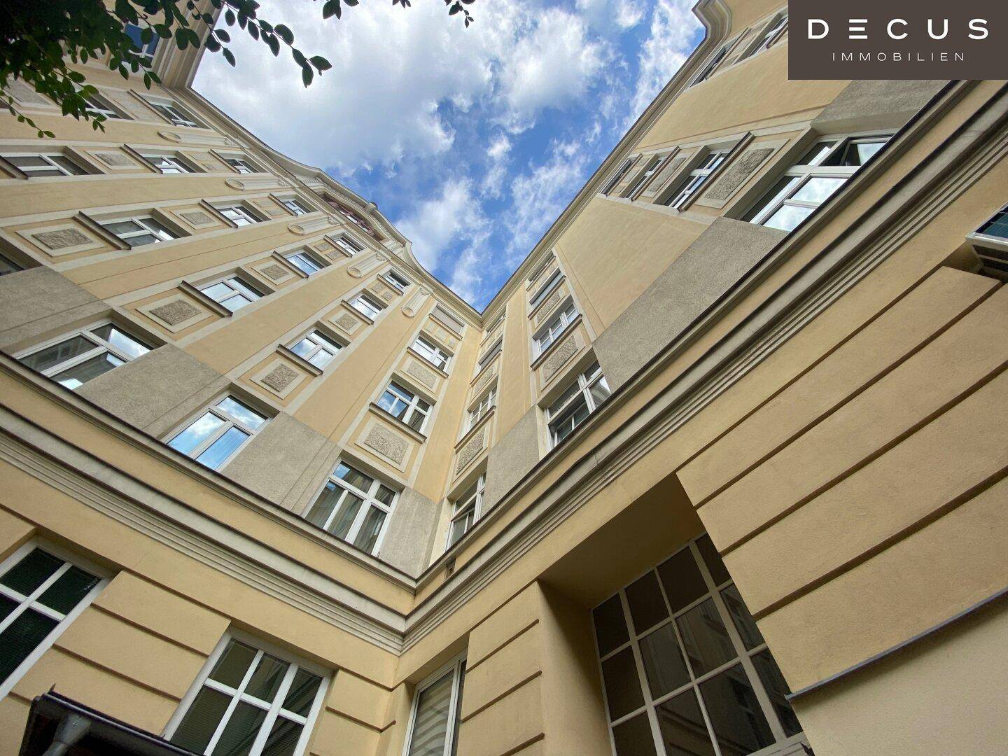 Blick nach oben auf die helle Fassade eines Altbaus mit vielen Fenstern und dekorativen Stuckelementen im Innenhof.
