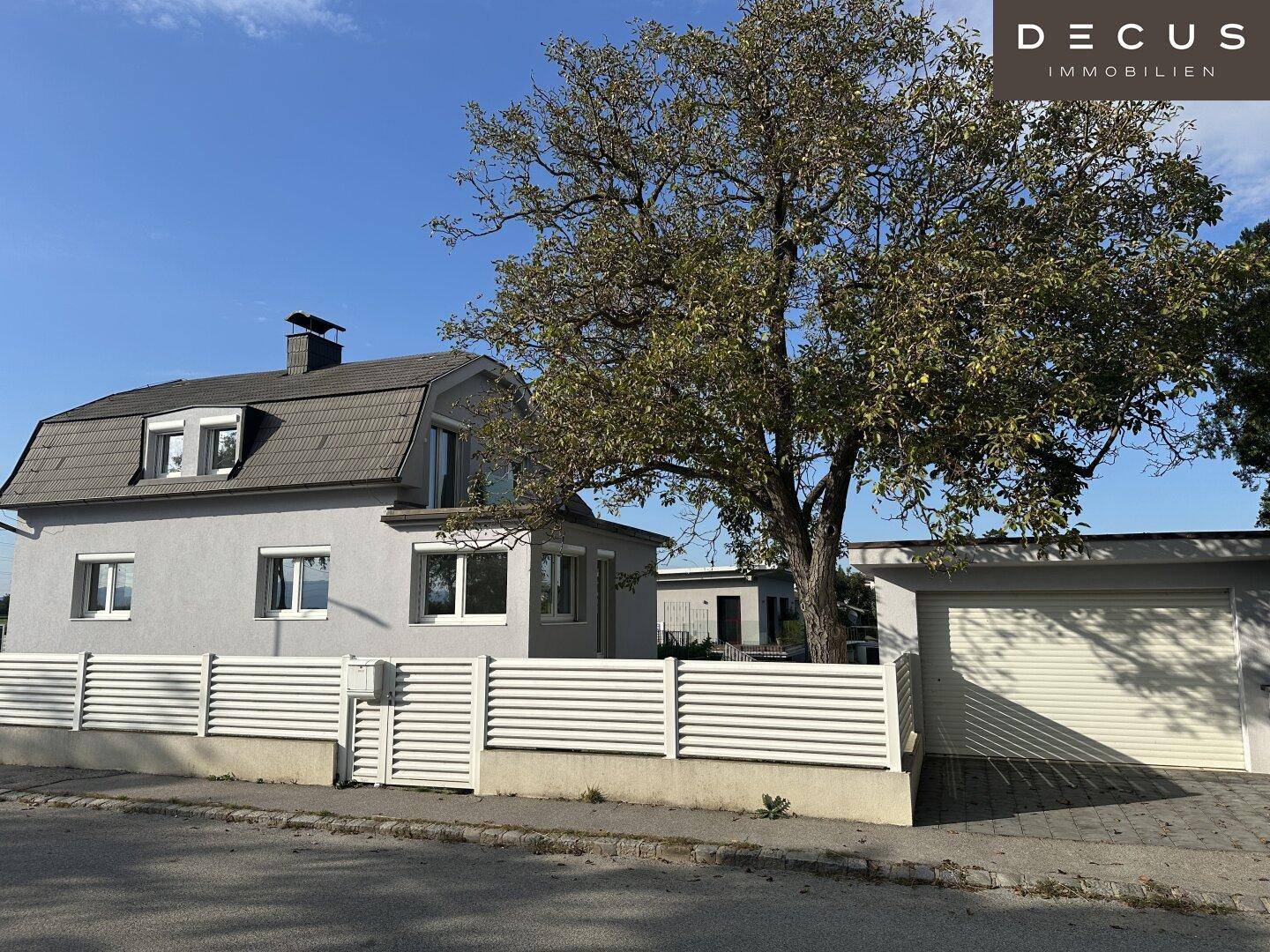 Ansicht des Hauses von der Straße mit Garage, Zaun und einem großen, alten Baum.
