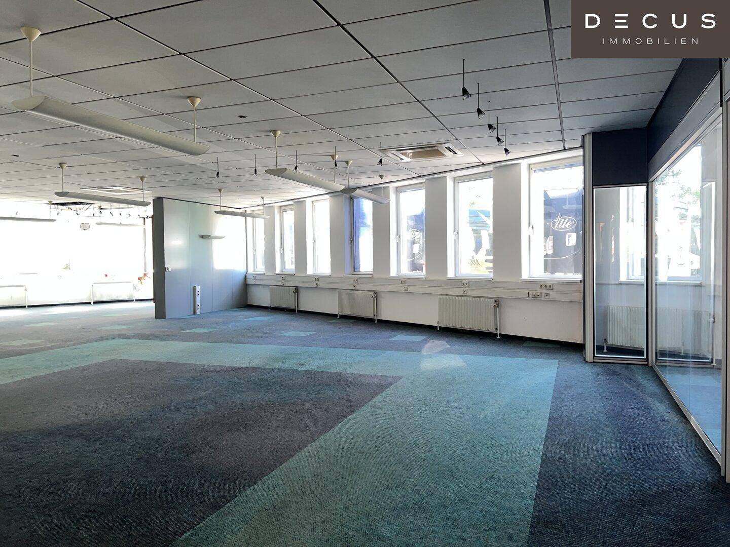 Geräumiger Bürobereich mit Teppichboden, großen Fenstern und einer modernen Glasabtrennung.
