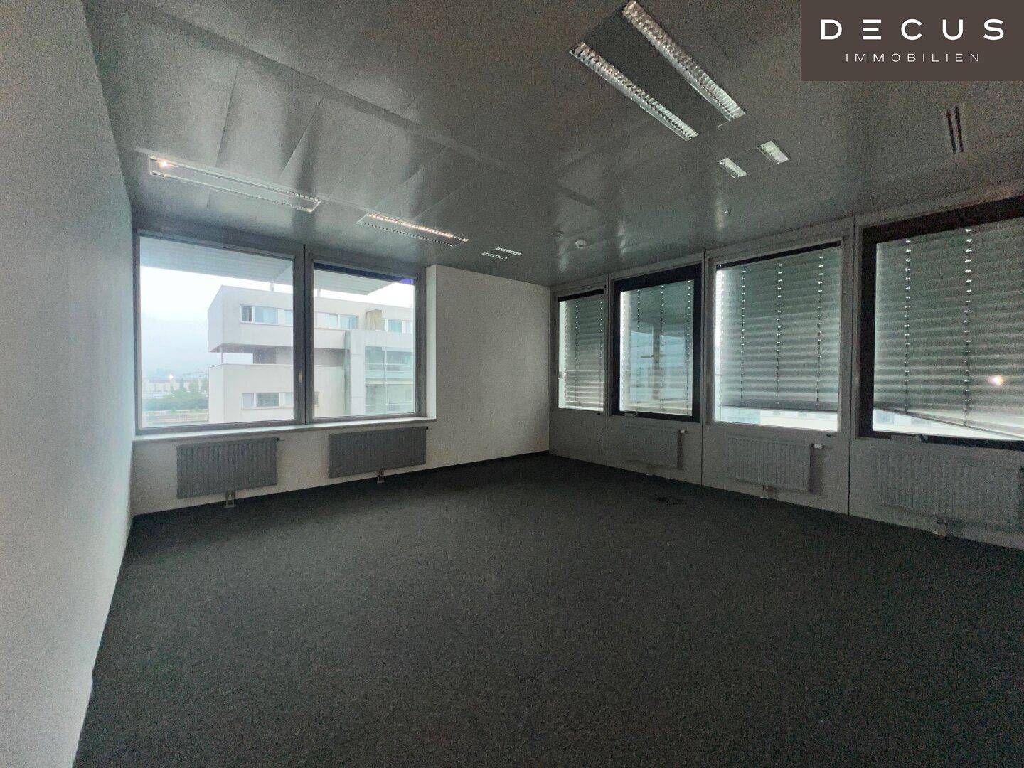 Heller Büroraum mit Teppichboden und Fenstern, die einen Blick auf umliegende Gebäude bieten.